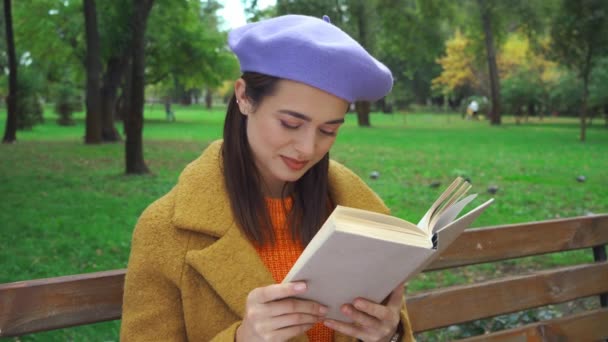 szczęśliwa kobieta siedzi w parku, czyta książkę, uśmiecha się i odwraca wzrok - Materiał filmowy, wideo