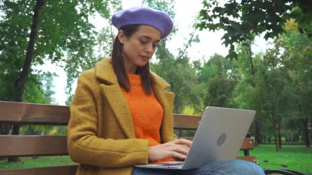 freelancer w stroju jesiennym wpisując na laptopie na ławce w parku - Materiał filmowy, wideo