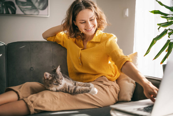 Casual woman in yellow shirt working on laptop with her cat on sofa, sitting together in room with window ang green plant - Photo, Image