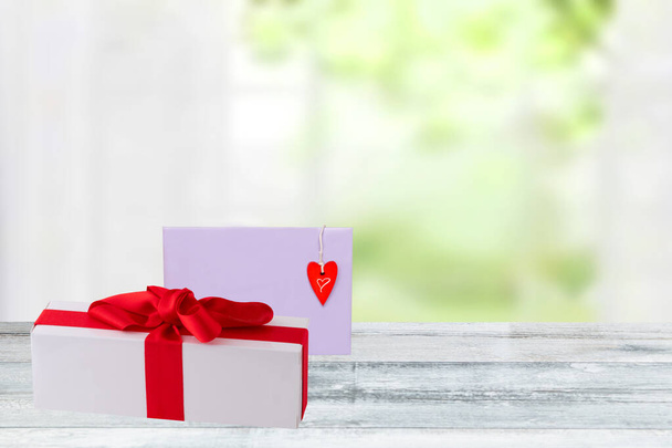 Valentines day background. Gift box with red ribbon and a letter with a red heart on a wooden tabletop over a blurred bright window background. Space for design. - Φωτογραφία, εικόνα