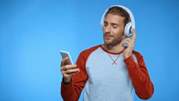 vzrušený muž s smartphonem tanec a zpěv ve sluchátkách izolovaných na modré - Záběry, video