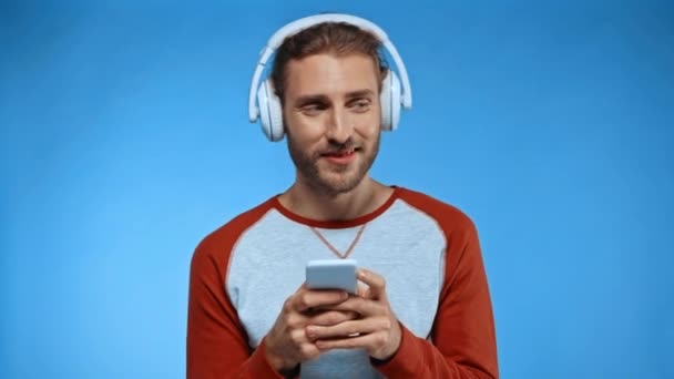 veselý muž ve sluchátkách zprávy na mobilním telefonu izolované na modré - Záběry, video