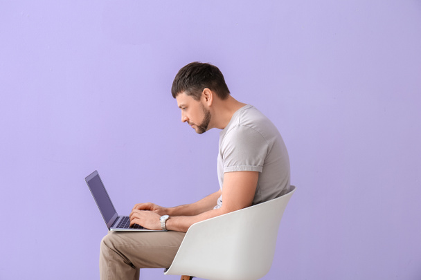 Mann mit schlechter Körperhaltung benutzt Laptop, während er auf Stuhl vor farbigem Hintergrund sitzt - Foto, Bild