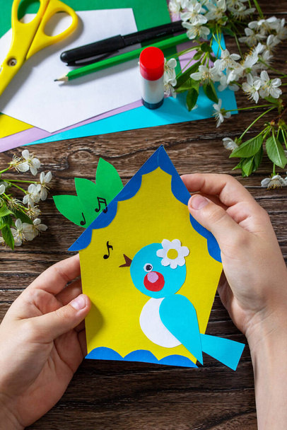 The child holds a spring postcard birdhouse with a bird on a wooden table. Handmade. Project of children's creativity, handicrafts, crafts for kids. - Photo, Image