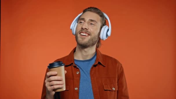 potěšený muž ve sluchátkách těší chuť kávy izolované na oranžové - Záběry, video