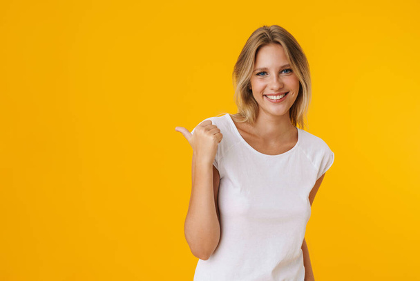 Cheerful beautiful girl smiling while pointing finger aside isolated over yellow background - Fotó, kép
