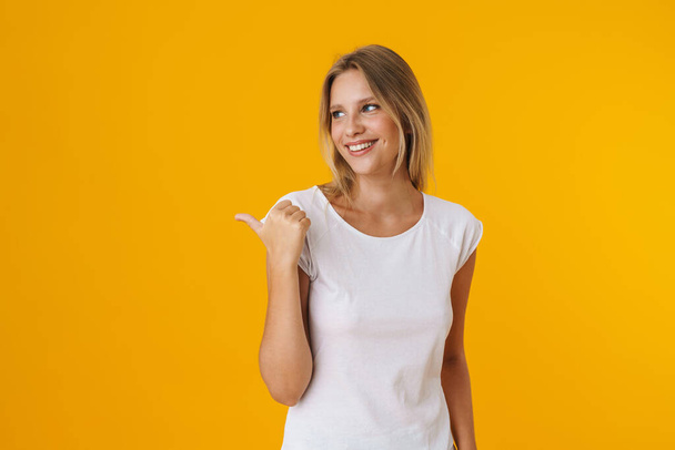 Cheerful beautiful girl smiling while pointing finger aside isolated over yellow background - Fotoğraf, Görsel