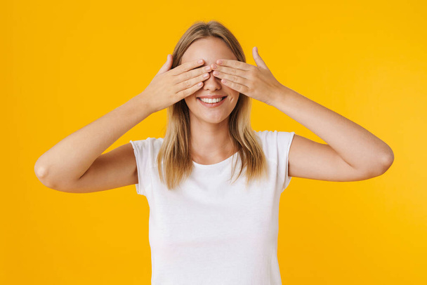Cheerful beautiful girl smiling while covering her eyes isolated over yellow background - Foto, afbeelding
