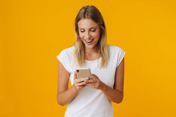Izgalmas szőke lány mosolyog, és használja a mobiltelefon elszigetelt sárga háttér - Fotó, kép