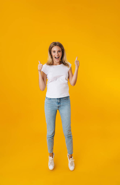 Excited beautiful girl jumping and showing thumbs up isolated over yellow background - Fotografie, Obrázek