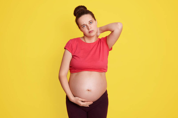 Pregnant woman standing with pain in neck, keeping hand on her bare tummy, looking directly at camera, has hair bun, wearing red t shirt, feels pain, being tired. - Фото, изображение