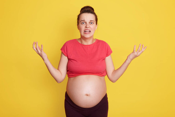 Young beautiful pregnant female with shocked facial expression, lady expecting baby, stands isolated over yellow background, being afraid, surprise, feels panic, spreading hand aside. - Foto, Imagem
