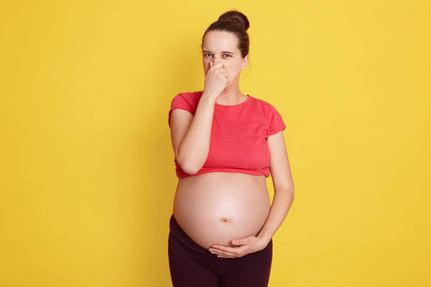 Young beautiful Caucasian pregnant woman expecting baby, stands isolated over yellow background, smelling something stinky and disgusting, intolerable smell, holding breath with fingers on nose. - Zdjęcie, obraz
