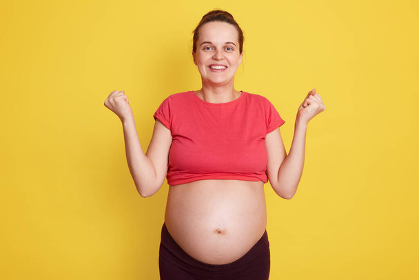 Pregnancy, motherhood, expectation baby. Close up portrait of happy pregnant woman with big bare belly standing with excitement expression isolated over yellow background, keeping fists clenched. - Photo, Image