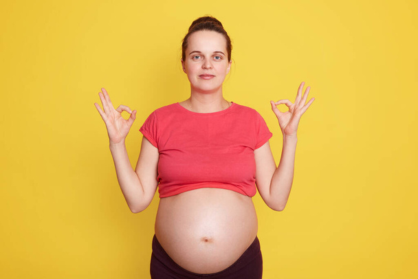Successful pregnancy. Positive Caucasian girl showing ok sign with both hands, lady with hair bun wearing casual t shirt, shows her bare belly, standing isolated over yellow background. - Фото, изображение
