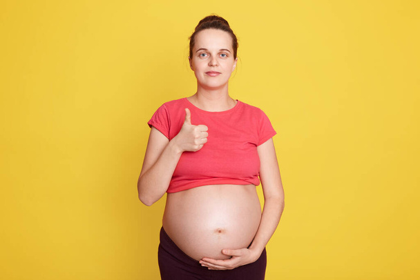 Beautiful European woman posing against yellow wall and showing thumb up, touching her bare tummy with hand, wearing red t shirt, has hair bun, expectant mother with positive emotions. - Photo, image