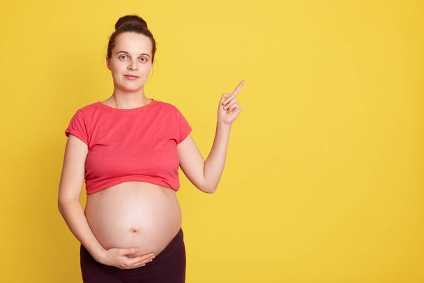 Caucasian young pretty pregnant woman touching belly and pointing aside isolated over yellow background, expectant mother with bare tummy wearing red t shirt. Copy space for advertisement. - Photo, image