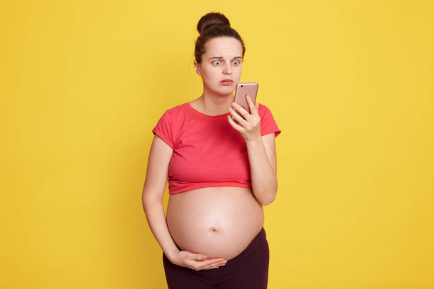 Caucasian pregnant female holds hand on belly and using smart mobile phone, preparing to be motherhood, looking at mobile phone screen with scared and surprised expression. - Fotoğraf, Görsel