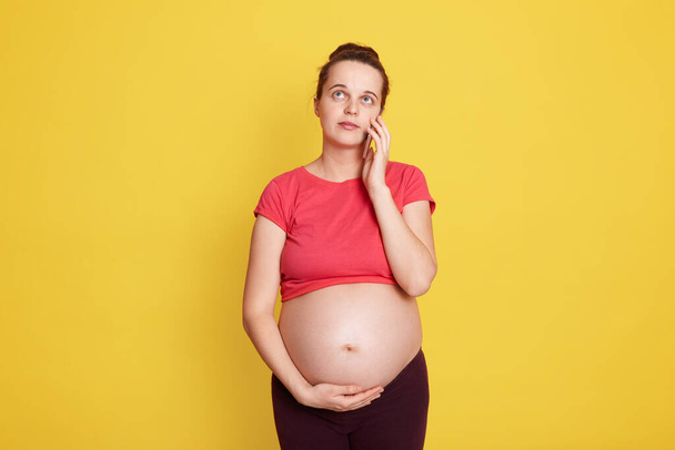 Smiling pregnant woman talking on her smart phone while posing isolated over yellow background, touching her bare tummy and looking away with dreamily expression. - Foto, Imagen