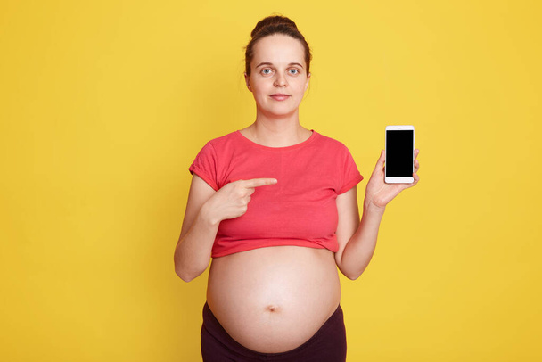 Mujer embarazada joven con aspecto agradable que muestra el teléfono con pantalla en blanco mientras está de pie aislado sobre la pared amarilla, futura madre con la barriga desnuda apuntando con el dedo delantero en el teléfono móvil. - Foto, imagen