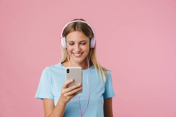 Vrolijk blond meisje in hoofdtelefoon met behulp van mobiele telefoon geïsoleerd over roze achtergrond - Foto, afbeelding