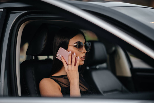 Jeune fille élégante assise dans une voiture de classe affaires dans une robe noire et parlant au téléphone. Mode et style d'affaires - Photo, image