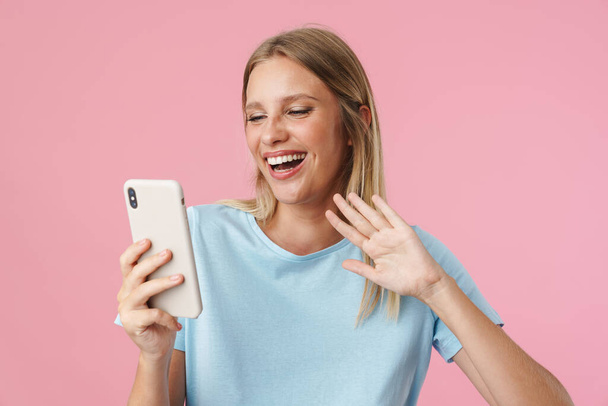 Cheerful girl waving hand while taking selfie photo on cellphone isolated over pink background - Fotó, kép