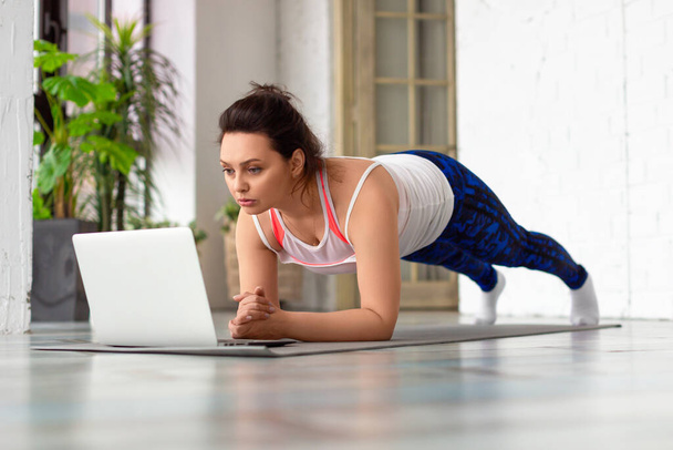 Žena středního věku cvičí doma online před notebookem. V pozici prkna. - Fotografie, Obrázek