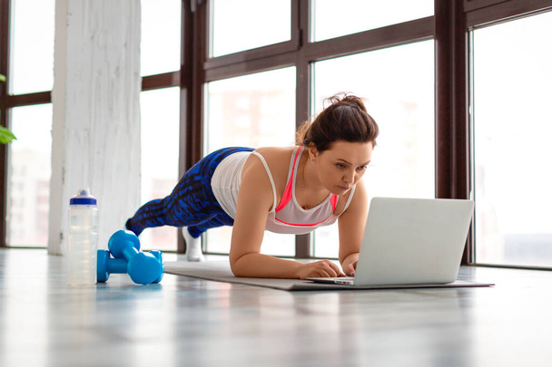 Mujer de mediana edad haciendo ejercicio en casa en línea delante de un monitor portátil. En posición de tablón. - Foto, imagen