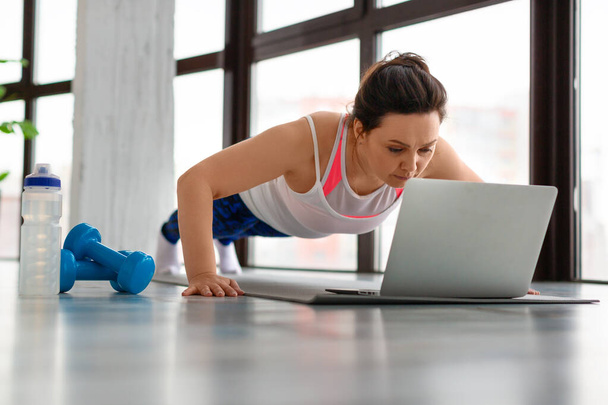 Middle-aged woman doing push-ups at home in front of a laptop monitor. - Фото, зображення
