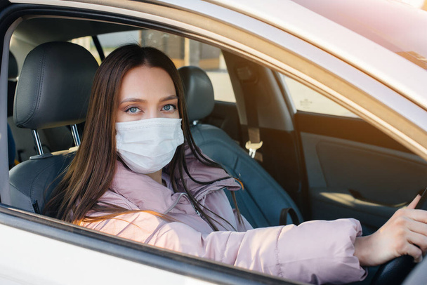 A young girl sits behind the wheel in the car in the mask during the global pandemic and coronavirus. Quarantine - Foto, imagen