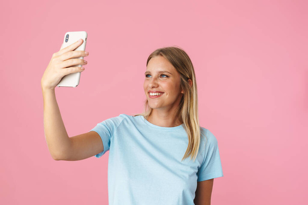 Cheerful beautiful girl smiling while taking selfie photo on cellphone isolated over pink background - Fotoğraf, Görsel