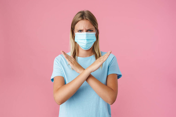 Blonde displeased girl in face mask showing stop gesture on camera isolated over pink background - Foto, imagen