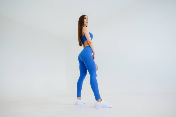 Sexy young girl performs exercises with a weight in a blue tracksuit on a white background. Fitness, healthy lifestyle - Fotó, kép