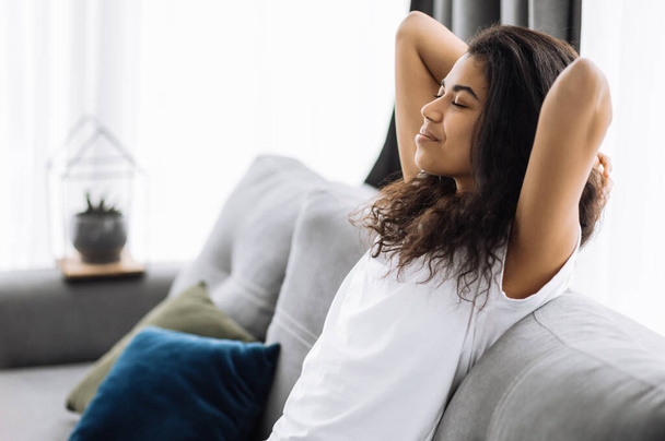 Une jeune femme afro-américaine satisfaite se relaxe sur le canapé dans le salon à la maison, souriant. Fille prend une pause de travail en ligne à distance, ou avoir des loisirs paresseux. Happy attrayant rêve féminin - Photo, image
