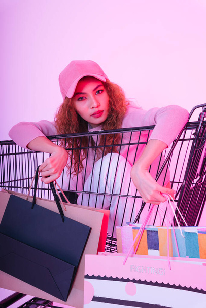Portrait of young asian curly hair girl sitting in shopping trolley with paper bags. - Zdjęcie, obraz
