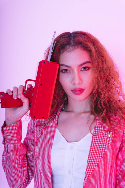 Portrait of young asian curly hair woman in pink jacket with red money gun on neon light background. - Photo, Image