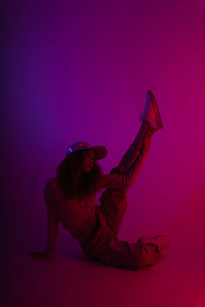 Αθλητική Ασιάτισσα φοράει καπέλο καθισμένη στο πάτωμα τεντώνοντας το πόδι σε μαύρο νέον φόντο. - Φωτογραφία, εικόνα