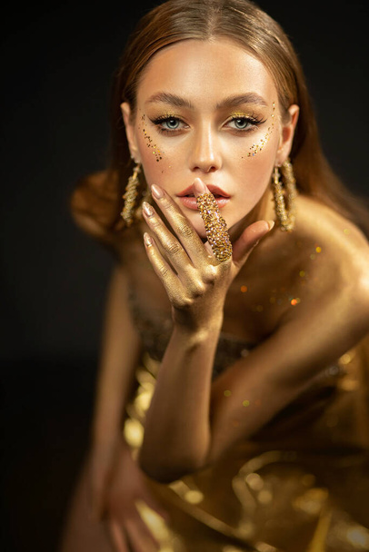 Young fashion model woman with golden body posing in studio. Background is black. Perfect makeup gold, glitter. Girl glamorous queen. Jewelry ring, earrings, accessories elegant dress. Beautiful face - Foto, Imagen