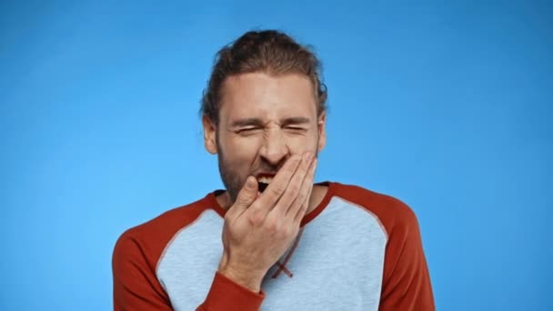 ospalý muž zívá a zakrývá ústa s rukou izolovanou na modré - Záběry, video