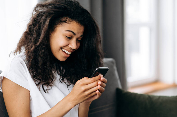 Teenager african american pretty girl sits at the couch in living room and uses her smartphone. Beautiful girl in casual wear spends time at home, chatting with friends or family, surfing internet - Valokuva, kuva