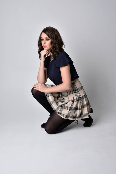 Full length portrait of pretty brunette woman wearing tartan skirt and blouse.  Sitting pose on the ground,  against a  studio background. - Photo, image