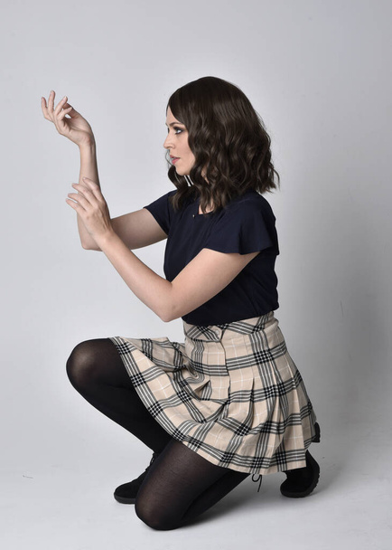 Full length portrait of pretty brunette woman wearing tartan skirt and blouse.  Sitting pose on the ground,  against a  studio background. - Fotoğraf, Görsel