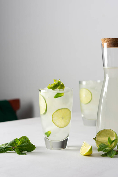 Освежающие холодные напитки с лаймом и мятой, стаканы с лимонадом и льдом, кувшин с напитком на белом фоне - Фото, изображение