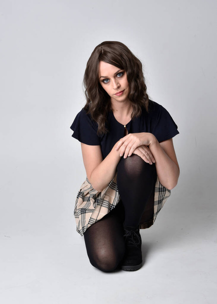 Full length portrait of pretty brunette woman wearing tartan skirt and blouse.  Sitting pose on the ground,  against a  studio background. - Photo, Image
