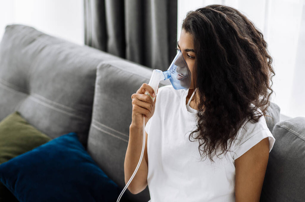 Nahaufnahme einer kranken afrikanisch-amerikanischen Frau mit einem Inhalator. Ungesundes Millennial-Mädchen beim Inhalieren zu Hause, sie verwendet Vernebler und Inhalator für die Behandlung sitzend auf der Couch zu Hause in Quarantäne - Foto, Bild