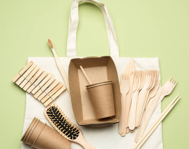 white textile bag and disposable tableware from brown craft paper on a green background. View from above, plastic rejection concept, zero waste  - Фото, зображення