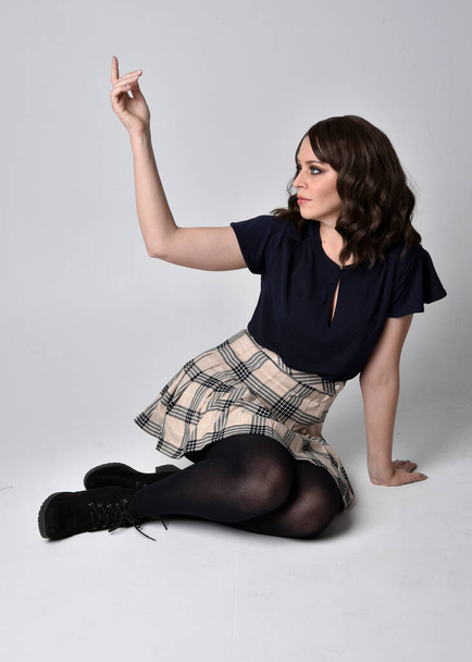 Full length portrait of pretty brunette woman wearing tartan skirt and blouse.  Sitting pose on the ground,  against a  studio background. - Photo, Image