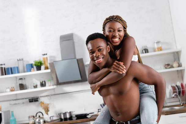 Musculoso africano americano hombre piggybacking alegre novia en sujetador en cocina  - Foto, imagen