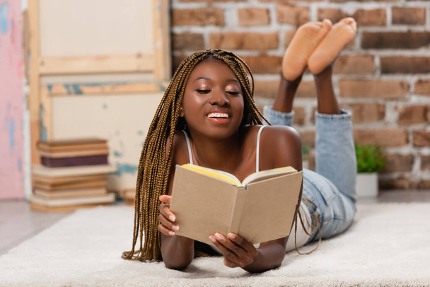 Sorridente donna africana americana leggere libro mentre si trova sul tappeto a casa - Foto, immagini